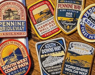 A series of Adventure Patch Company patches arrayed on a wooden table. These brightly-coloured embroidered cloth patches feature the trail name, distance, start and end points, and a distinctive central image to represent the trail.