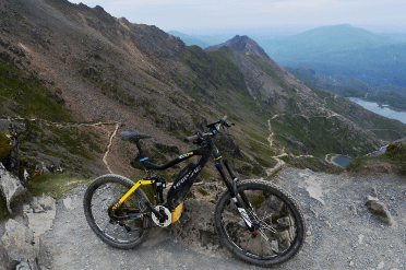 A bicycle stands propped against a rock at the top of Snowdon, with the valley behind.