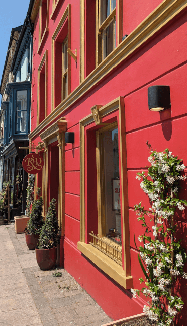 Red shoe shop in Machynlleth. This terraced shop, one of many along Mach's pretty high street, is painted vibrant red with gold accents and decorative plants outside.